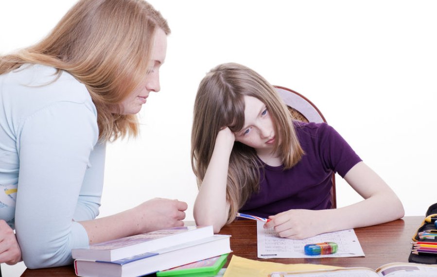Ein Mädchen im Schulalter sitzt bei den Hausaufgaben am Schreibtisch.  Auf dem Tisch liegen verschiedene Arbeitsmaterialien. Sie stützt ihren Kopf mit dem Arm ab und wirkt überfordert. Ihre Mutter beugt sich zu ihr herüber. 