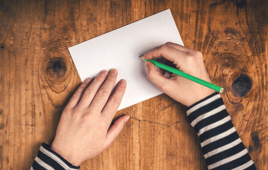 Auf einer Holztischplatte liegt ein Briefumschlag. Die eine Hand einer Person hält einen Stift und die andere Hand liegt auf dem Briefumschlag.