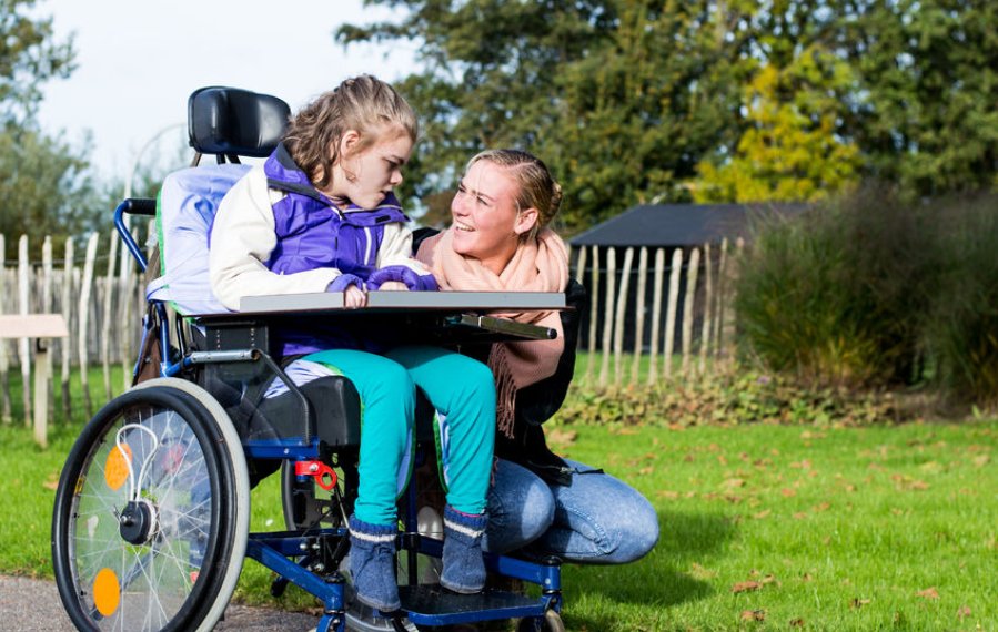 Offene Hilfen - Mädchen in einem Rollstuhl ist in einem Garten. Neben ihr kniet eine junge Frau. Diese lacht das Mädchen an.