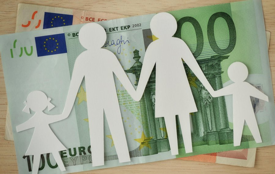 Auf einem Tisch liegen zwei Geldscheine. Ein fünfzig Euro und ein hundert Euro Schein liegen übereinander. Auf den beiden Geldscheinen wiederum liegt eine Silhouette aus Papier. Die Silhouette stellt von links nach rechts ein Mädchen, einen Mann, eine Frau und einen Jungen dar. Sie halten sich an der Hand und symbolisieren eine Familie.