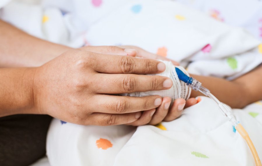 Die Hände eines Erwachsenen halten die Hand eines Kindes in einem Krankenhausbett. An der Hand des Kindes ist ein Zugang gelegt.