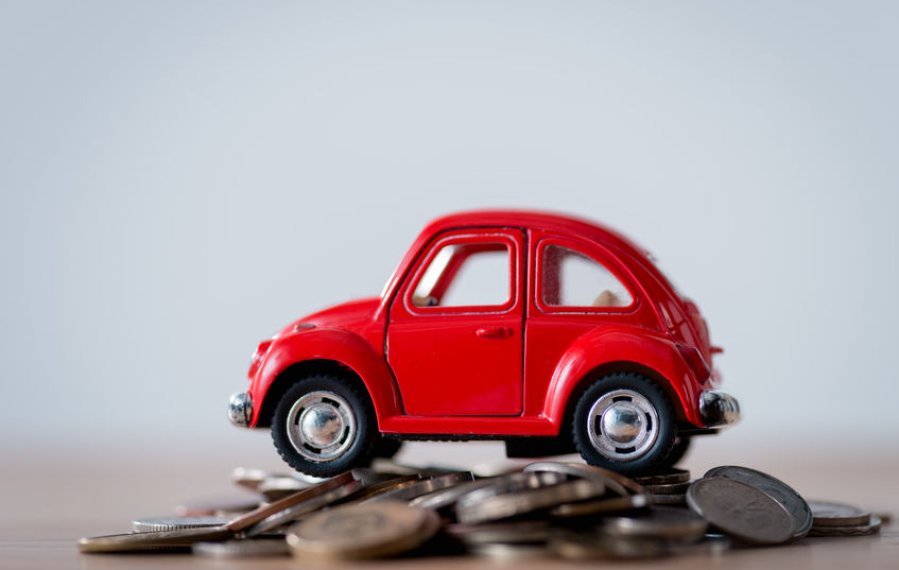 Ein kleines rotes Spielzeugauto steht auf einem Berg mit Geldmünzen. Die Münzen liegen auf einem Tisch.