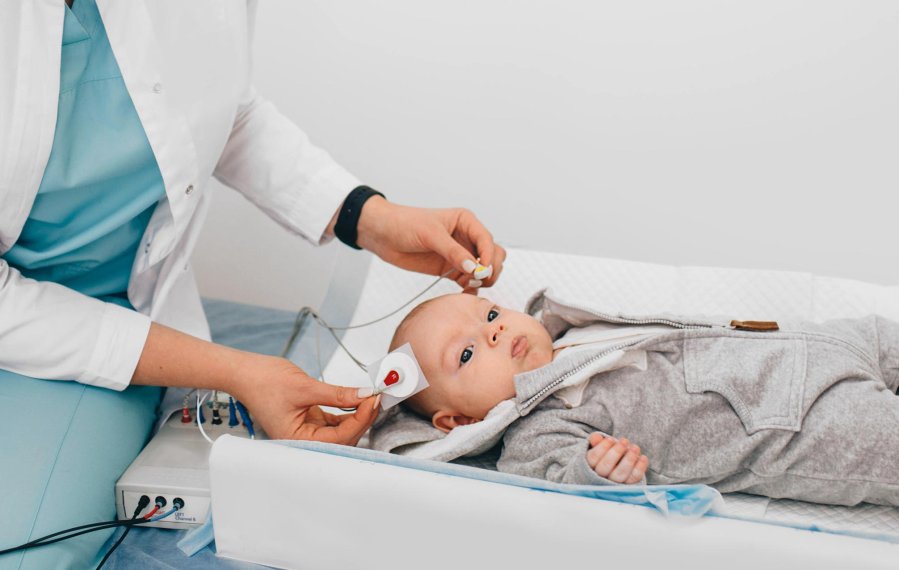 Ein Baby liegt auf einer weißen Unterlage. Dem Baby werden von einer Ärztin Elektroden am Kopf befestigt.