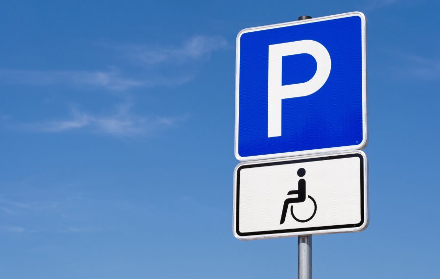 Ein blaues Verkehrsschild mit einem großen weißen P darauf. Darunter hängt ein weißes Verkehrsschild, auf welchem ein Rollstuhlfahrer abgebildet ist. Im Hintergrund ist blauer Himmel.