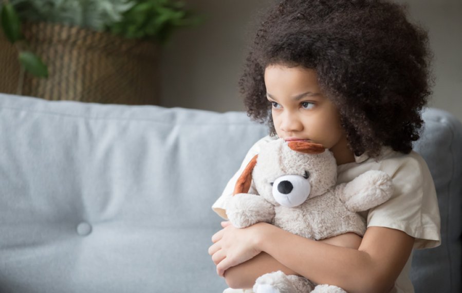 Ein kleines Mädchen sitzt auf der Couch. Sie hält einen Teddybär fest im Arm und schaut ins Leere. Sie wirkt abwesend.