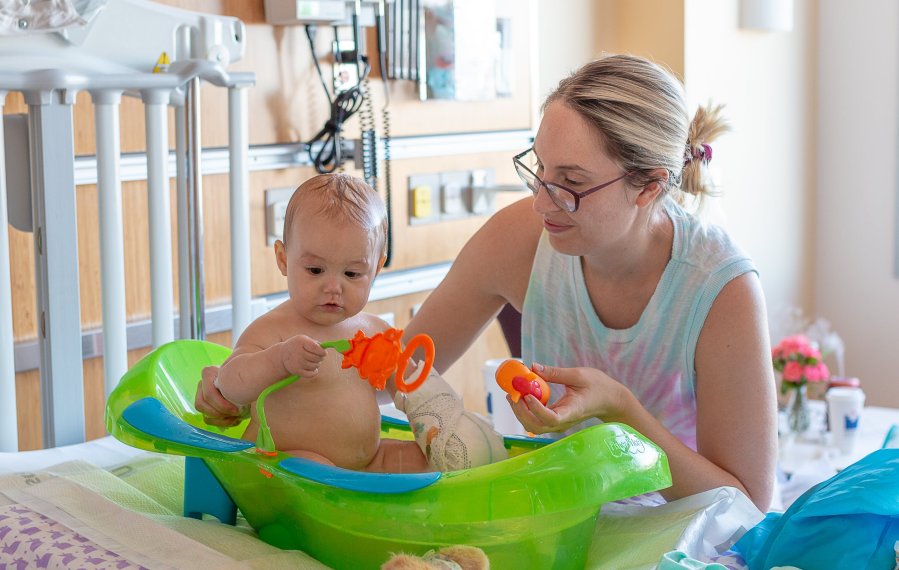 Eine Mutter badet ihr Baby in einer Wanne auf einem Krankenbett. Das Baby hat einen Verband am linken Arm.