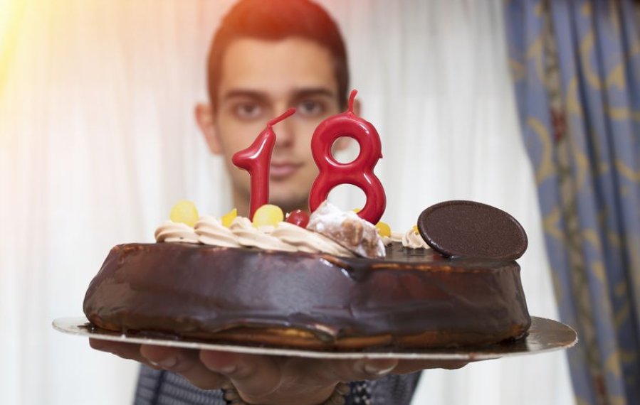 Ein junger Mann hält eine Torte in die Kamera. Darauf stecken rote Kerzen in der Zahlenform 18.