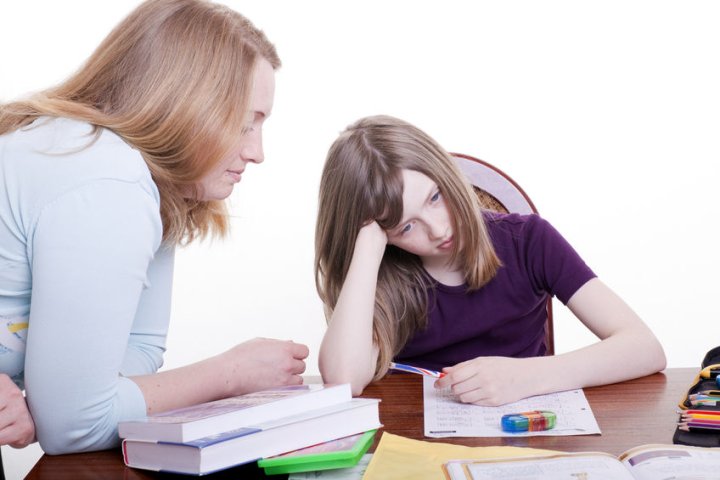 Diagnose ADHS - Ein Mädchen im Schulalter sitzt bei den Hausaufgaben am Schreibtisch.  Auf dem Tisch liegen verschiedene Arbeitsmaterialien. Sie stützt ihren Kopf mit dem Arm ab und wirkt überfordert. Ihre Mutter beugt sich zu ihr herüber.