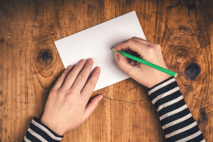 Auf einer Holztischplatte liegt ein Briefumschlag. Die eine Hand einer Person hält einen Stift und die andere Hand liegt auf dem Briefumschlag.