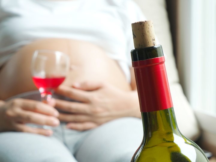 Diagnose Fetales Alkoholsyndrom - eine Schwangere liegt auf dem Sofa, zu sehen ist der Babybauch. In der Hand hält sie ein gefülltes Weinglas. Im Vordergrund steht die Rotweinflasche.