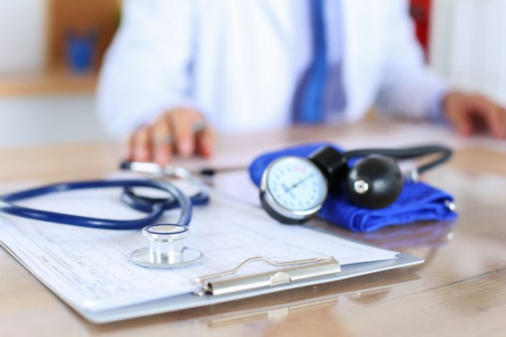 Gesetzliche Krankenversicherung - auf einem Tisch liegen ein Stethoskop, ein Blutdruckmessgerät und eine Klemmbrett mit Papieren. Im Hintergrund ist ein Arzt zu sehen.