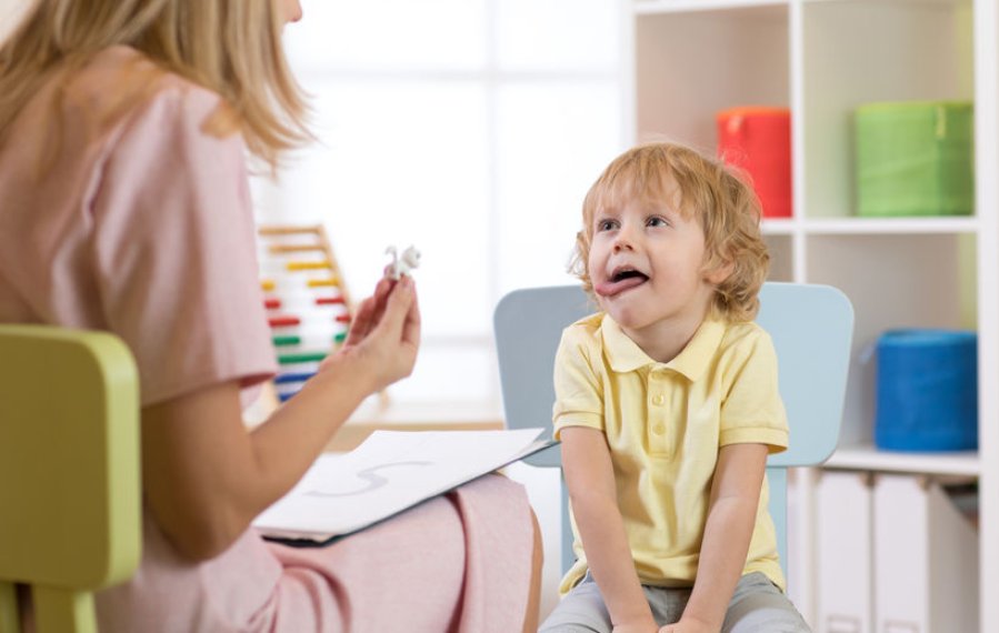 Ein Junge im Kindergartenalter sitzt auf einem Stuhl. Er streckt die Zunge aus dem Mund und hält sich mit den Händen am Stuhl fest. Er wirkt interessiert und konzentriert. Ihm gegenüber sitzt eine Frau. Auf ihrem Schoß liegt ein Blatt Papier. Auf diesem ist ein großes S abgebildet. Die Frau hält eine kleine Figur in ihrer Hand.