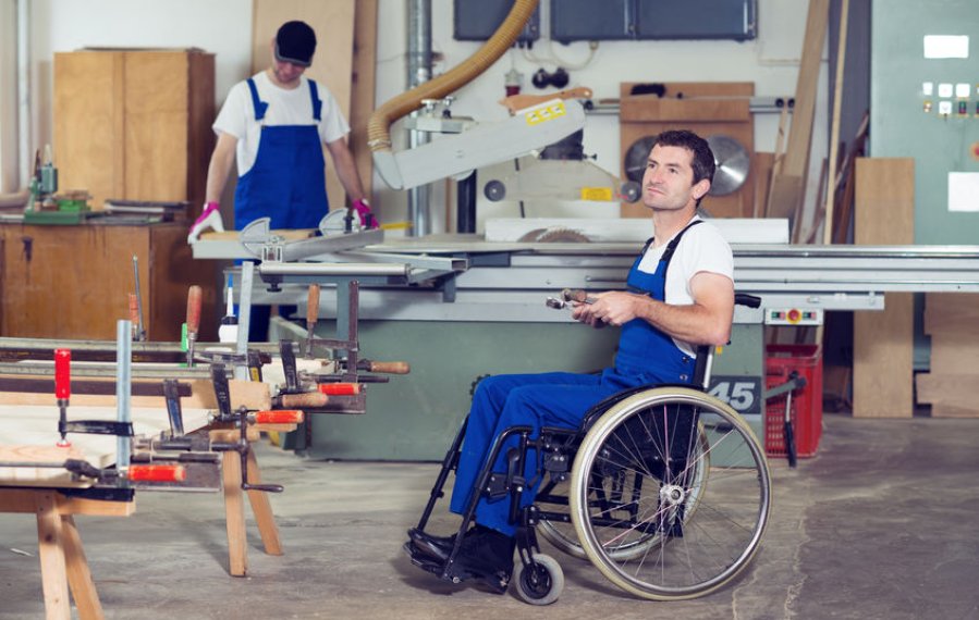 Ein junger Rollstuhlfahrer im Blaumann in einer Schreinerei-Werkstatt. Im Hintergrund bedient ein weiterer Mann eine Maschine.