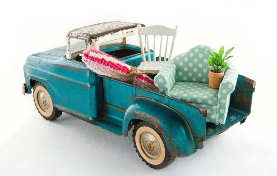 Ein türkises Spielzeugauto, welches auf der Laderampe einen Teppich, einen Stuhl, einen Sessel und eine Pflanze geladen hat.