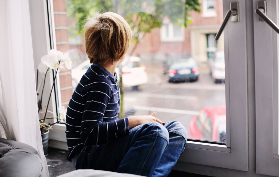 Diagnose Autismus - Ein Junge sitzt auf dem Fensterbrett des Wohnzimmerfensters und schaut hinaus auf die Straße