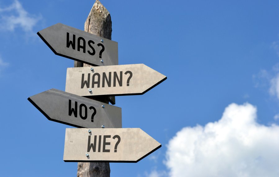 Das Bild zeigt einen Wegweiser aus Holz vor blauem Himmel. Die Fragen „Was?“, „Wann?“, „Wo?“ und „Wie?“ zeigen jeweils in eine andere Himmelsrichtung.