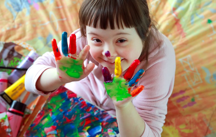 Ein junges Mädchen sitzt auf dem Boden. Auf ihrem Schoß liegt ein bunt angemaltes Papier. Darauf liegen Farbtuben. Sie hält beide Hände mit ausgestreckten Finger nach oben und schaut lächelnd in die Kamera. Ihre Hände sind auch bunt angemalt. 