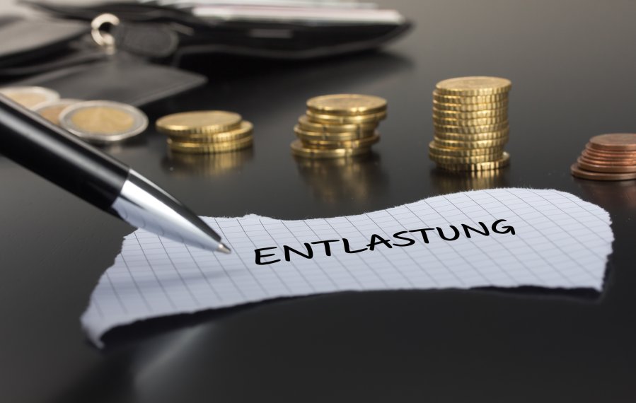 Ein Stück abgerissenes Papier liegt auf einem schwarzen Tisch. Auf dem Zettel steht das Wort „Entlastung“. Links im Bild ist ein Teil eines Kugelschreibers. Im Hintergrund liegen Euromünzen und ein Geldbeutel.