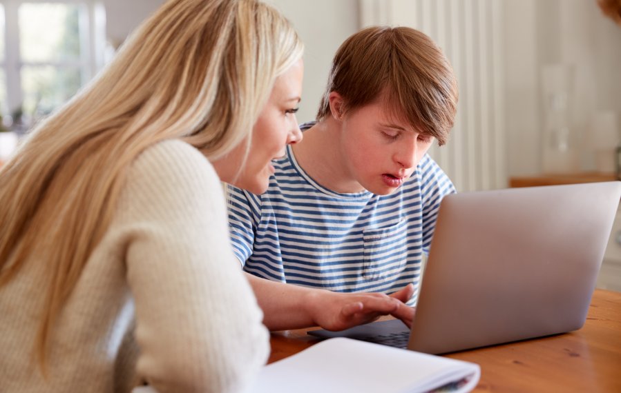 Erwachsenenbildung - Ein junger Mann sitzt an einem Holztisch. Vor ihm auf dem Tisch steht ein Laptop. Er tippt etwas auf der Tastatur. Neben ihm sitzt eine Frau mit langen blonden Haaren. Vor ihr liegt ein Block. 