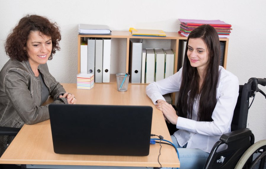 Zwei Frauen sitzen in einem Büro an einem Tisch. Eine der beiden Frauen sitzt im Rollstuhl. Auf dem Schreibtisch steht ein Laptop. Im Hintergrund stehen Ordner in einem Regal. 