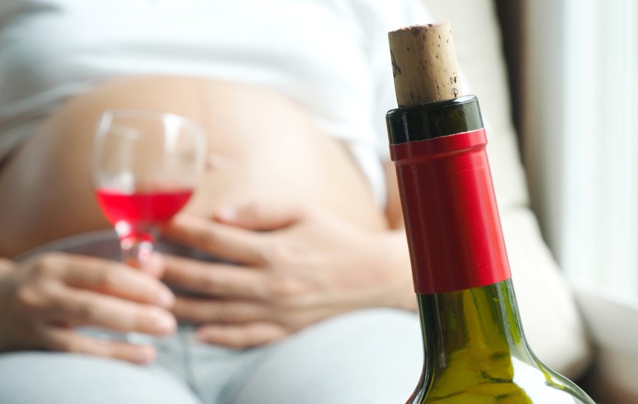 Eine Schwangere liegt auf dem Sofa, zu sehen ist der Babybauch. In der Hand hält sie ein gefülltes Weinglas. Im Vordergrund steht die Rotweinflasche.