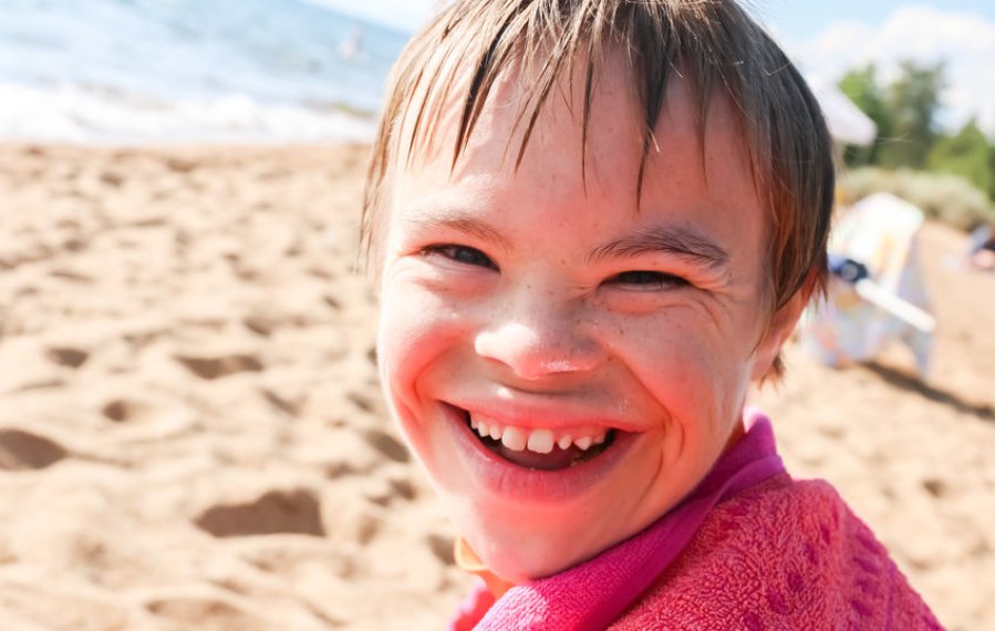 Ein Junge lacht in die Kamera. Seine Haare sind nass. Im Hintergrund sind der Strand und das Meer zu sehen.