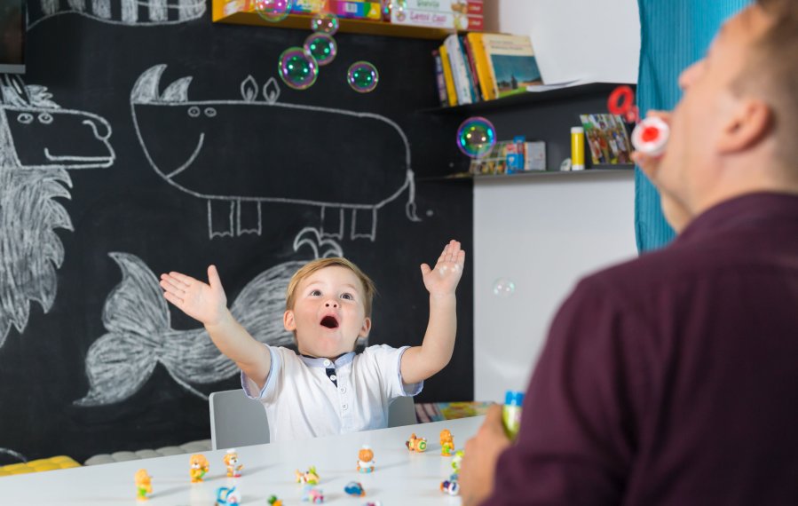 Ein kleiner Junge sitzt an einem Tisch. Im Hintergrund sind Spielsachen und eine Tafel. Auf dem Tisch stehen kleine bunte Figuren. Im Gegenüber sitzt ein Mann und macht Seifenblasen. Der Junge wirkt begeistert und streckt die Arme nach oben aus.