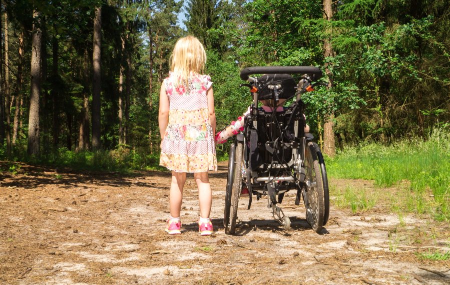 Ein kleines Mädchen und ein Kind im Rollstuhl befinden sich auf einem Weg im Wald. Die Sonne scheint. Sie halten sich an der Hand. 