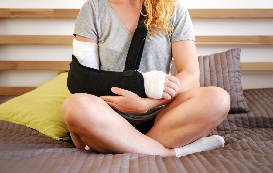 Ein Frau sitzt im Schneidersitz auf einem Bett. Ihr rechter Arm ist eingegipst und in einer Schlinge.