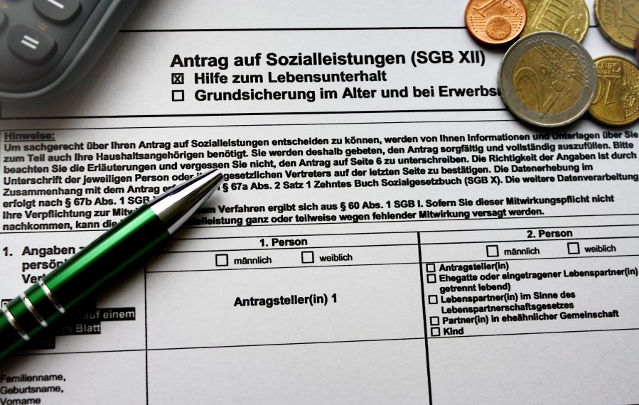 Ein Antrag auf Sozialleistungen bei dem die Hilfe zum Lebensunterhalt angekreuzt ist. Auf dem Antrag liegt ein Taschenrechner, ein Kugelschreiber und Geldmünzen.