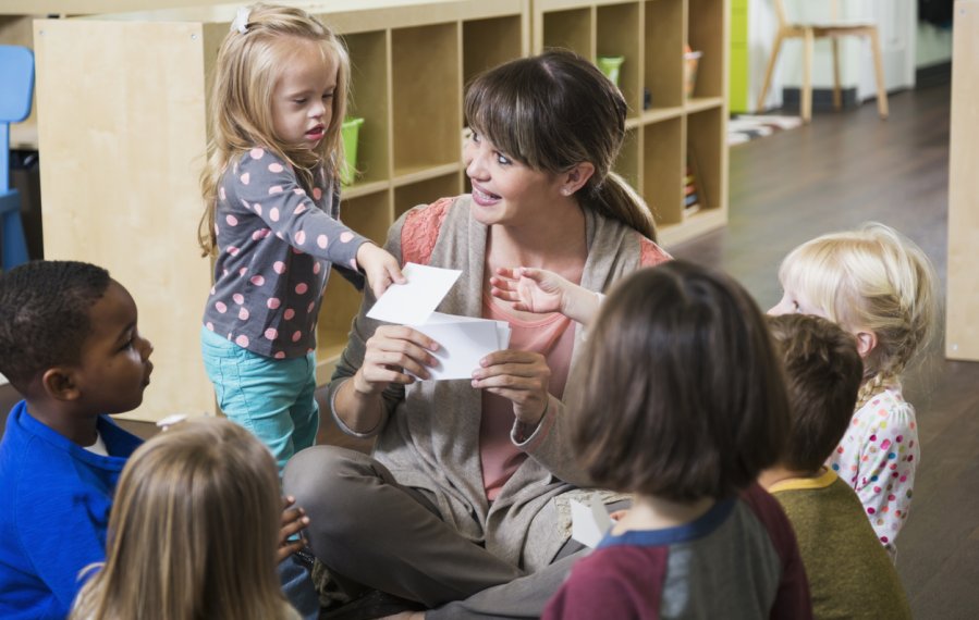 Eine kleine Gruppe von Kindern sitzen mit einer Erzieherin im Kreis. Die Erzieherin sammelt von allen Kindern einen Zettel ein.