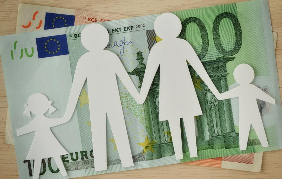 Auf einem Tisch liegen zwei Geldscheine. Ein fünfzig Euro und ein hundert Euro Schein liegen übereinander. Auf den beiden Geldscheinen wiederum liegt eine Silhouette aus Papier. Die Silhouette stellt von links nach rechts ein Mädchen, einen Mann, eine Frau und einen Jungen dar. Sie halten sich an der Hand und symbolisieren eine Familie.