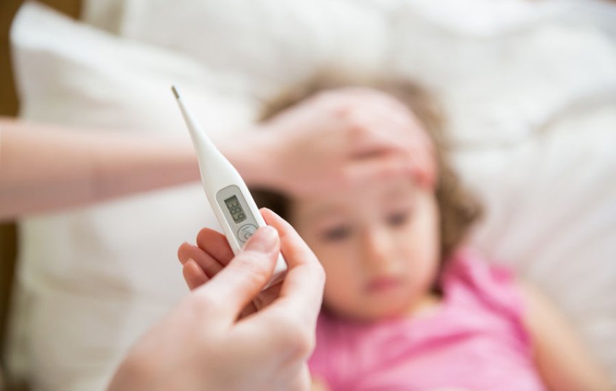 Eine Mutter hält ein Fieberthermometer in der Hand. Die andere Hand liegt auf der Stirn ihrer im Bett liegenden Tochter.