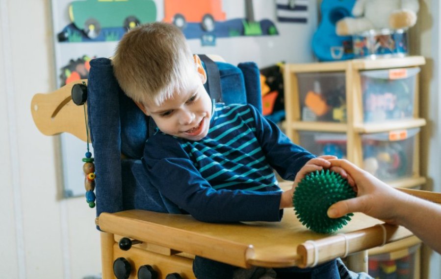 Ein Junge sitzt in einem Therapiestuhl. Sein Blick ist nach unten gesenkt. Er lächelt. Seine rechte Hand liegt auf einem grünen Igelball, der vor ihm auf dem Stuhltisch liegt. Der Igelball wird von einer weiteren Hand eines Erwachsenen gehalten. Im Hintergrund sind Bilder und Spielzeug zu sehen.
