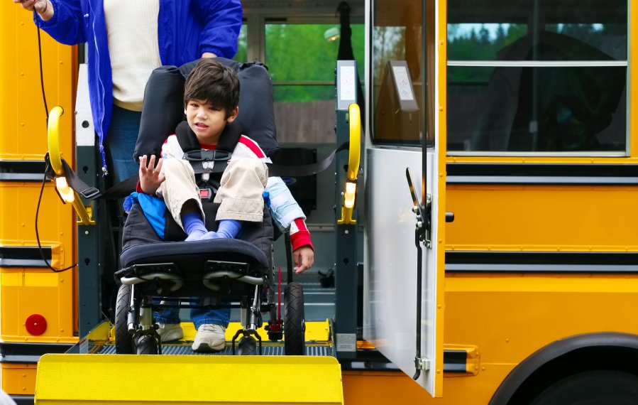 Ein Junge wird in seinem Rollstuhl über eine Rampe in einen gelben Bus befördert. Er lacht.