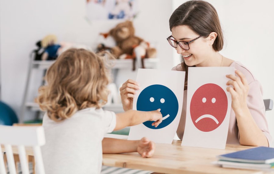 Ein kleines Kind und eine junge Frau sitzen sich an einem Tisch gegenüber. Die Frau hält in jeder Hand ein Blatt Papier. Auf dem einen ist ein großer blauer lachender Smiley und auf dem anderen ein großer roter trauriger Smiley abgebildet. Das Kind zeigt auf den lachenden Smiley.