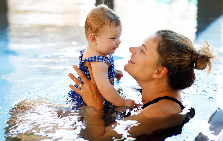 Eine Mutter ist mit ihrem Baby in einem Schwimmbecken. Sie hält das Baby vor sich und lacht es an.