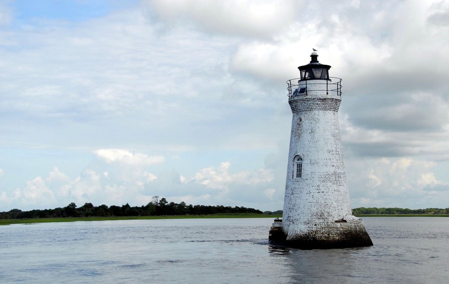 Ein weißer Leuchtturm steht im Wasser, in einer Bucht. Im Hintergrund sind Bäume auf dem Land.