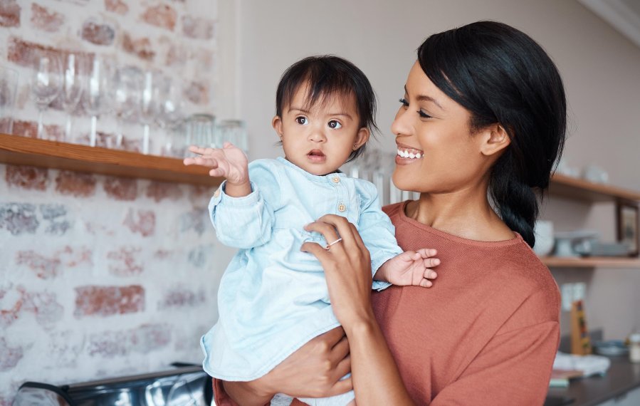 Eine glückliche Mutter hält ihr Baby mit Down-Syndrom auf dem Arm. Sie befinden sich in einer Küche in einem Einfamilienhaus.
