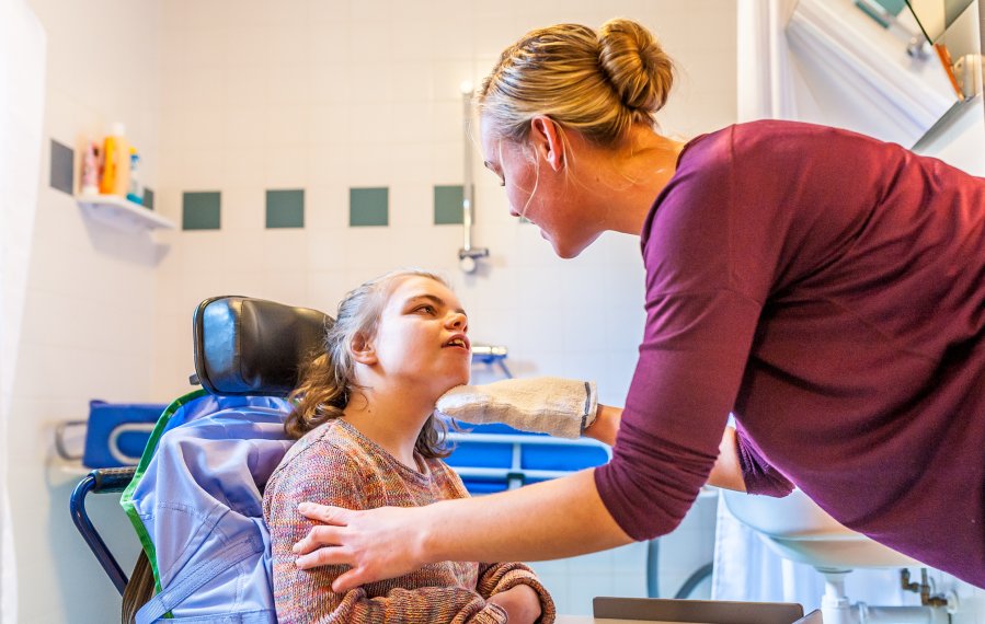 Zwei Frauen in einem Badezimmer. Die eine junge Frau sitzt im Rollstuhl. Die andere Frau beugt sich zu ihr und wäscht das Gesicht der jungen Frau mit einem Waschlappen. Die Frauen schauen sich gegenseitig in die Augen.
