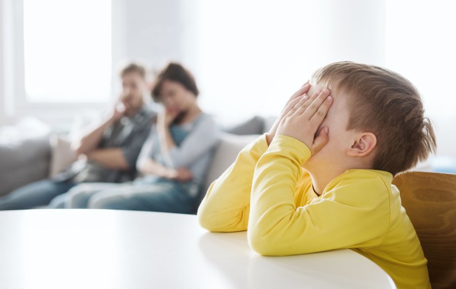 Ein Junge in gelben Pullover sitzt an einem Tisch und stützt seinen Kopf in die Arme. Dabei bedeckt er mit den Händen sein Gesicht. Im Hintergrund sieht man seine Eltern auf dem Sofa sitzen. Sie sehen nachdenklich aus.