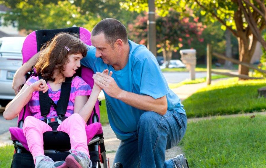 Ein Vater kniet neben seiner Tochter in einem lilafarbenen Rollstuhl auf dem Gehweg. Die Tochter hat die Augen geschlossen und den Kopf in die Richtung ihres Vaters gedreht. Der Vater schaut das Mädchen an, hält ihre Hand und hat einen Arm um sie gelegt.