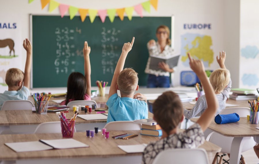 Unterstützungsangebote im Schulalter - Grundschüler sitzen in einem Klassenzimmer. Vor der Klasse steht eine Lehrerin. Sie zeigt auf einen Schüler um ihn aufzurufen. Hinter der Lehrerin ist eine Tafel. Auf dieser stehen Rechenaufgaben.