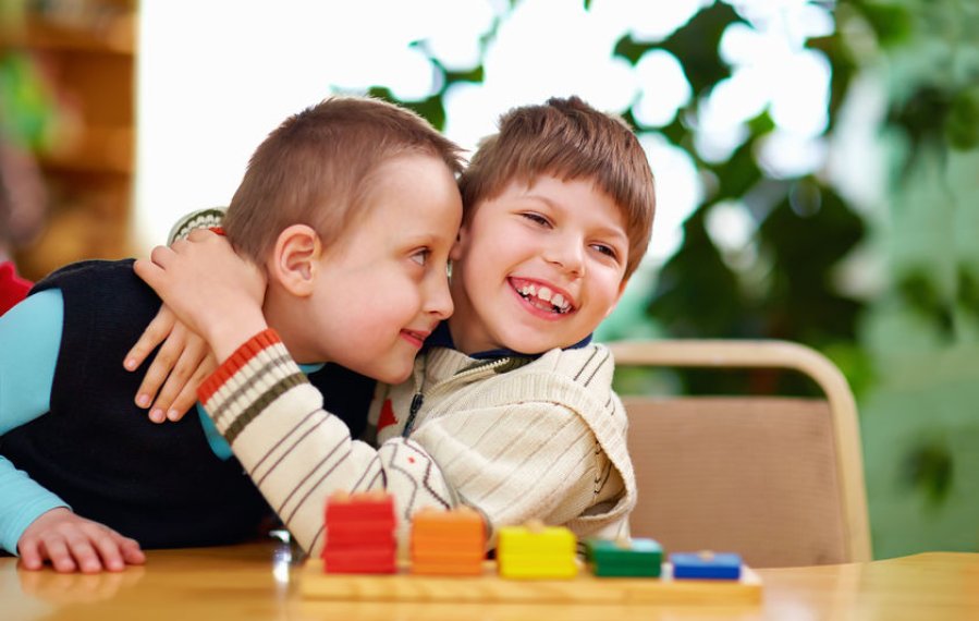Zwei Jungen im Grundschulalter sitzen an einem Tisch. Der Junge auf der rechten Seite des Bildes umarmt den anderen Jungen. Beide lachen. Vor ihnen steht ein Spiel mit bunten Holzplättchen.
