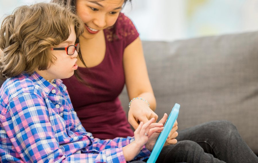 Ein Junge sitzt neben seiner Mutter auf dem Sofa. In der Hand hält er ein Tablet und bedient es. Die Mutter deutet darauf.
