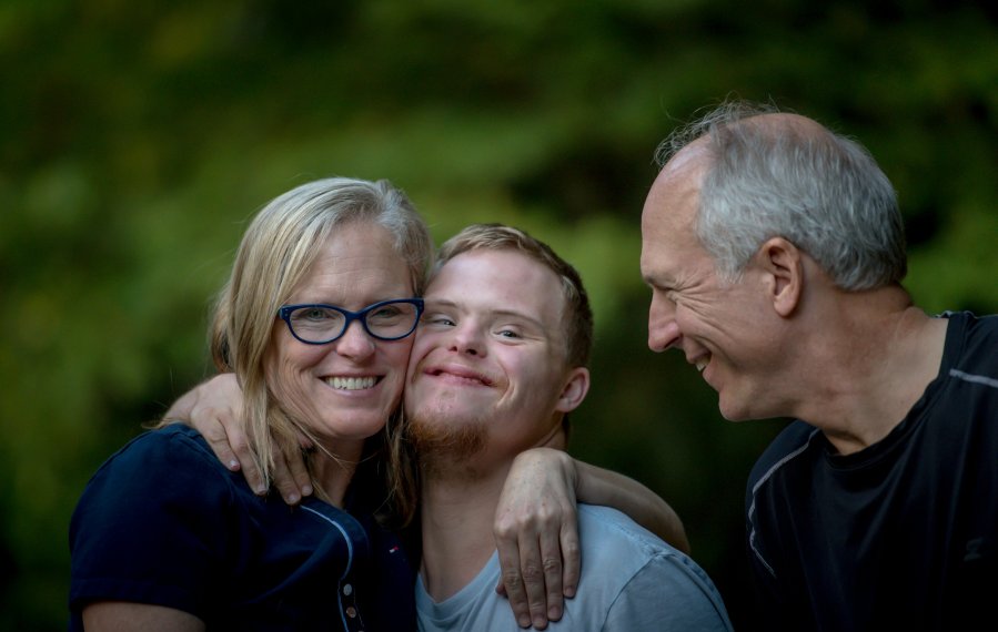 Eine Mutter umarmt ihren Sohn. Beide schauen in die Kamera. Der Vater sieht beide an und lächelt.