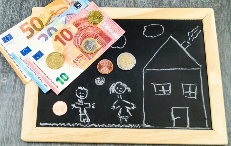 Eine kleine Tafel, auf der mit weißer Kreide ein Haus und zwei Kinder gemalt wurden. Darauf liegen Geldscheine und Münzen.