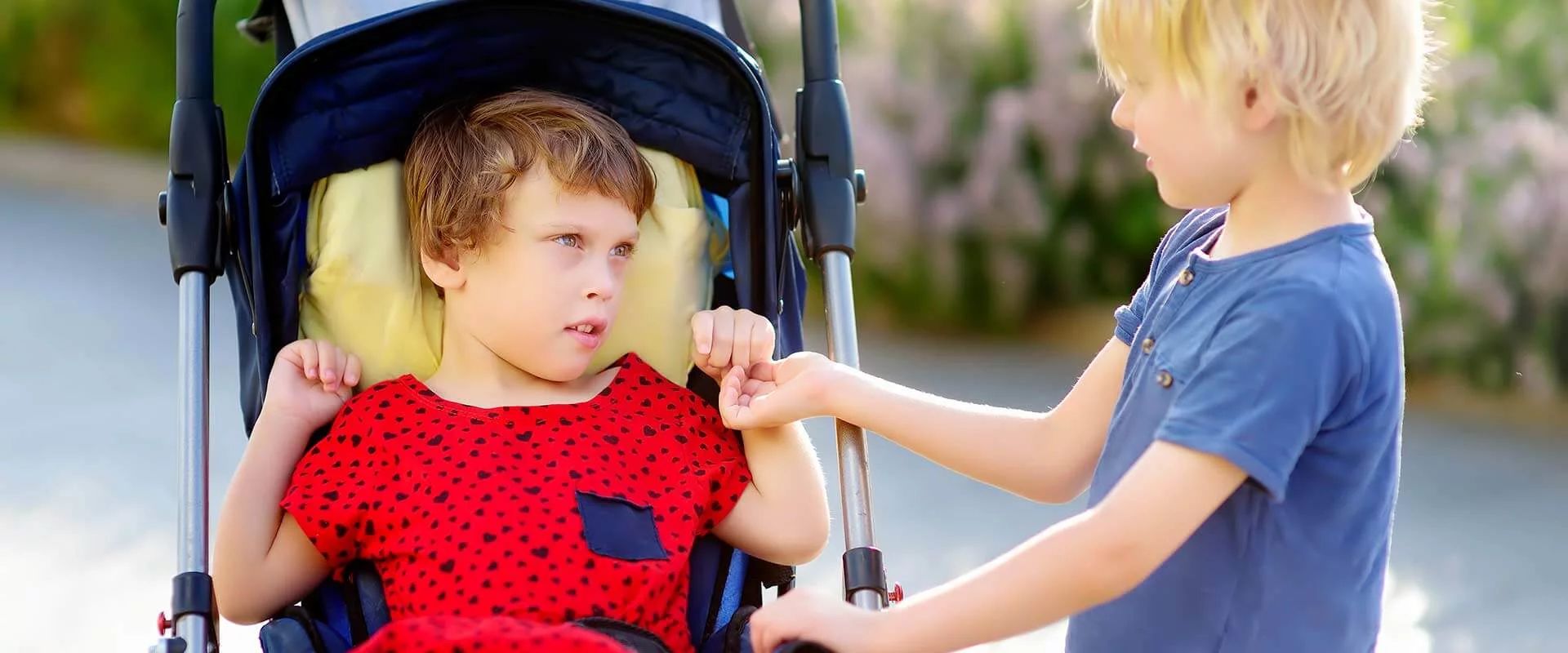 Ein Mädchen sitzt im Kinderwagen. Vor ihr steht ein kleiner Junge. Sie schaut diesen intensiv an. Der Junge hält eine Hand des Mädchens. Es ist Sommer.