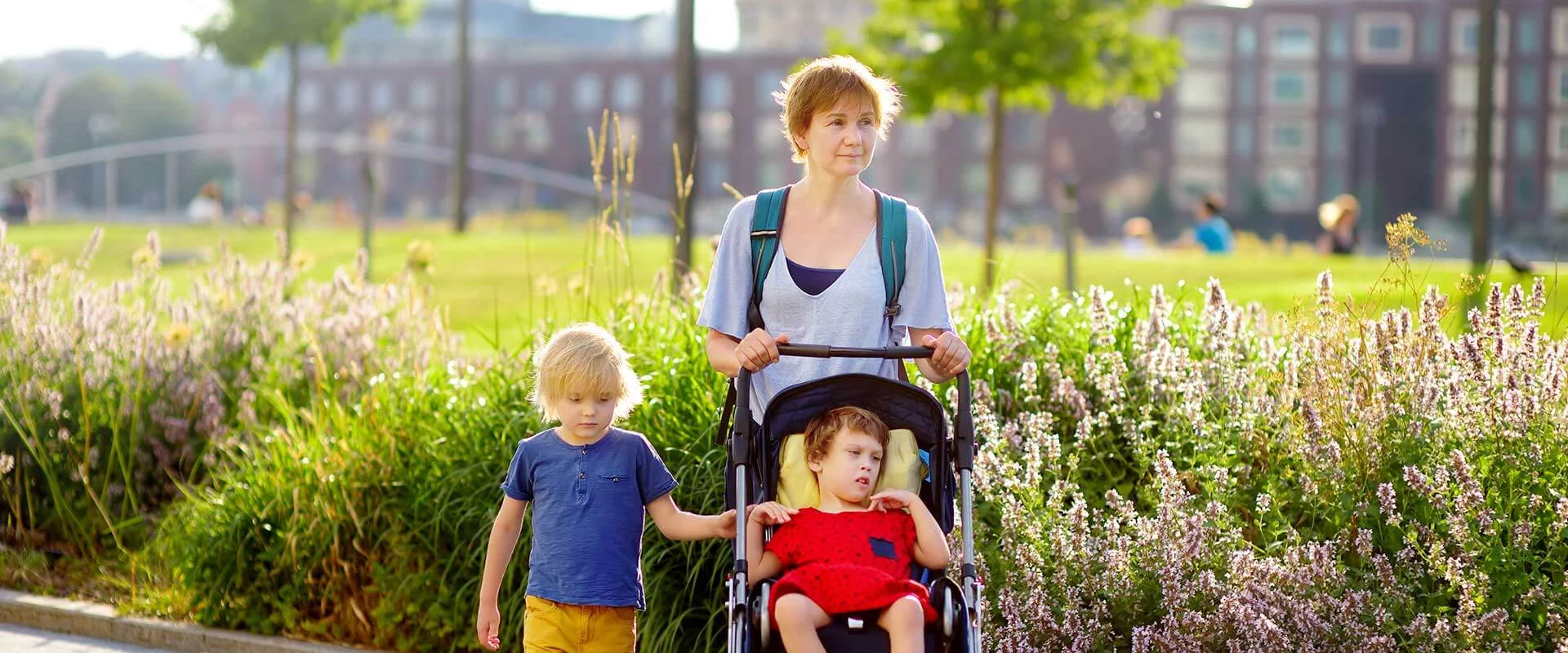 Eine Mutter geht mit ihren zwei Kindern spazieren. Das Mädchen sitzt im Kinderwagen, der Junge hält sich am Kinderwagen fest und läuft neben den beiden. Im Hintergrund sind Blumen und Bäume.